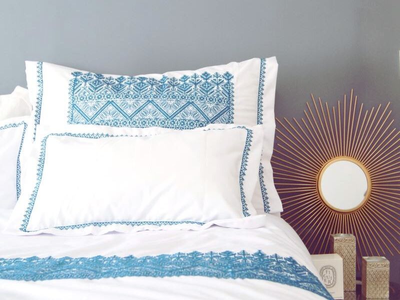 A close-up of decorative pillows featuring intricate blue Moroccan Fez embroidery on white fabric, accompanied by a modern gold sunburst mirror and patterned accessories in a stylish bedroom setting.