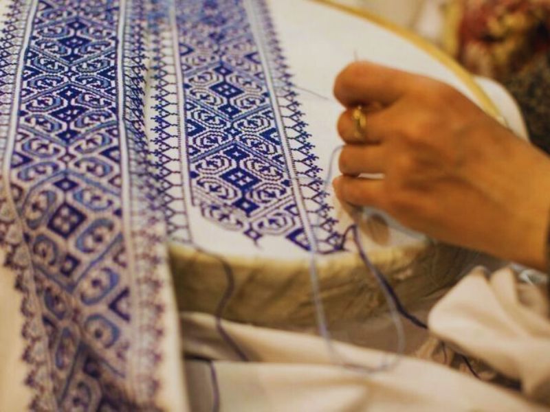 A close-up of a hand embroidering intricate blue patterns on white fabric, showcasing traditional Moroccan Fez embroidery.
