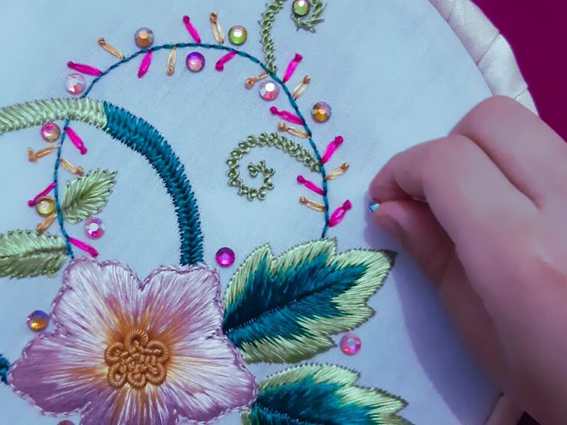 A close-up of a hand embroidering a floral design with colorful threads and sequins on a white fabric.