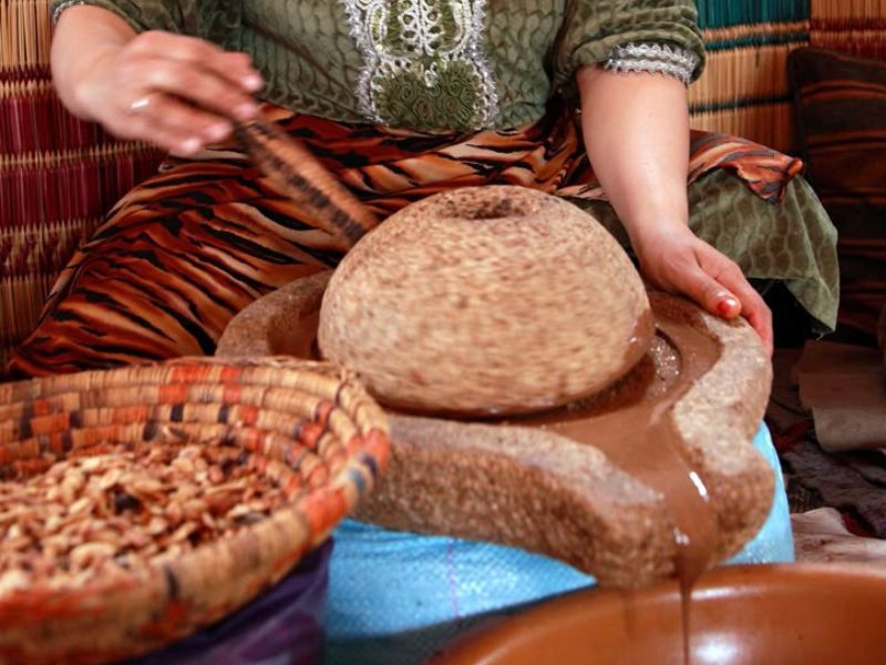 A woman grinding Argan nuts using a traditional stone grinder, with a woven basket and ground Argan paste visible.