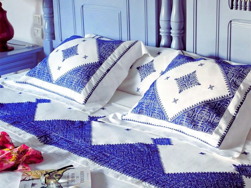 A beautifully embroidered Moroccan bedspread featuring intricate blue designs on white fabric, with matching pillows, placed on a stylish bed.