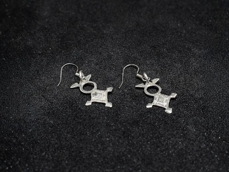 A pair of silver earrings shaped like the Agadez cross, featuring intricate designs and hanging hooks, displayed against a dark background.