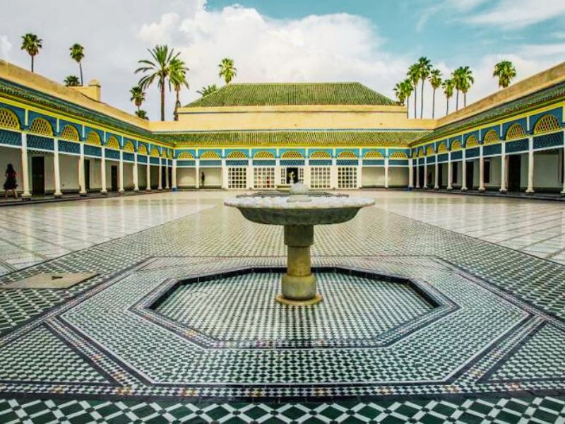 View of the Grand Courtyard of Bahia Palace, featuring intricate tile work and a central fountain, framed by palm trees and blue arches.