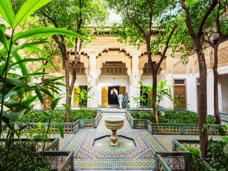 An interior courtyard of Bahia Palace in Marrakesh featuring lush greenery, decorative tile work, and a central fountain.