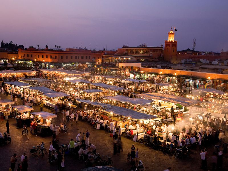 A vibrant evening scene in Jemaa el-Fna square, Marrakesh, filled with bustling food stalls and crowds, illuminated by warm lights against a dusky sky.