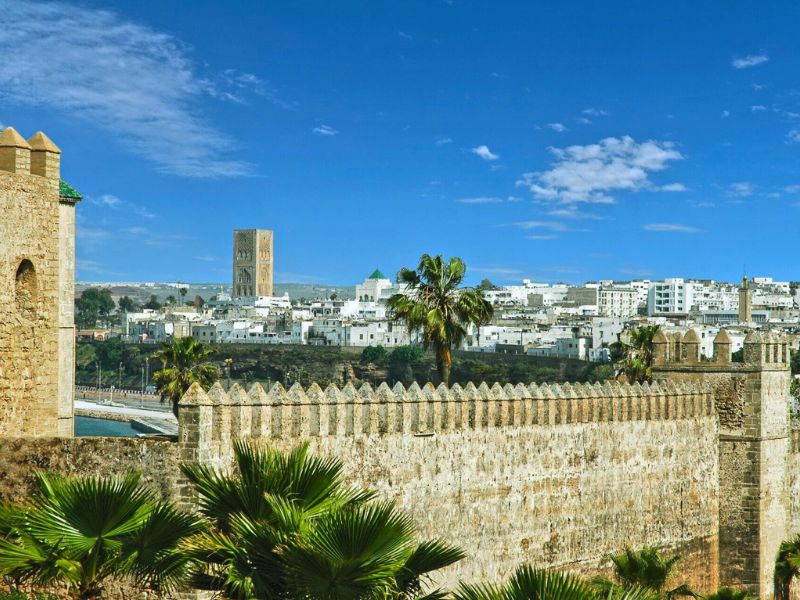 View of Rabat's historical skyline featuring a fortified wall, palm trees, and the Hassan Tower in the background under a clear blue sky.