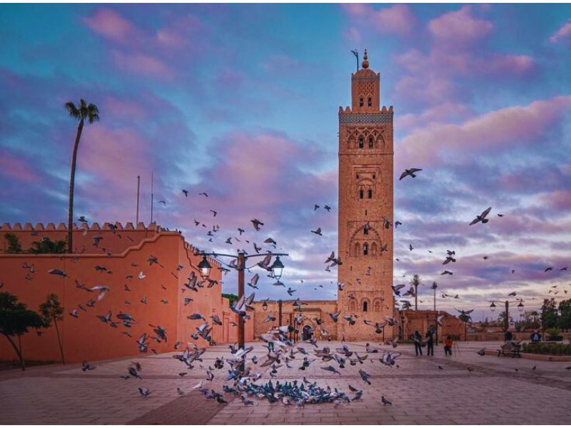 View of the Koutoubia Mosque in Marrakesh, with its towering minaret surrounded by flying pigeons and a colorful sky during sunset.