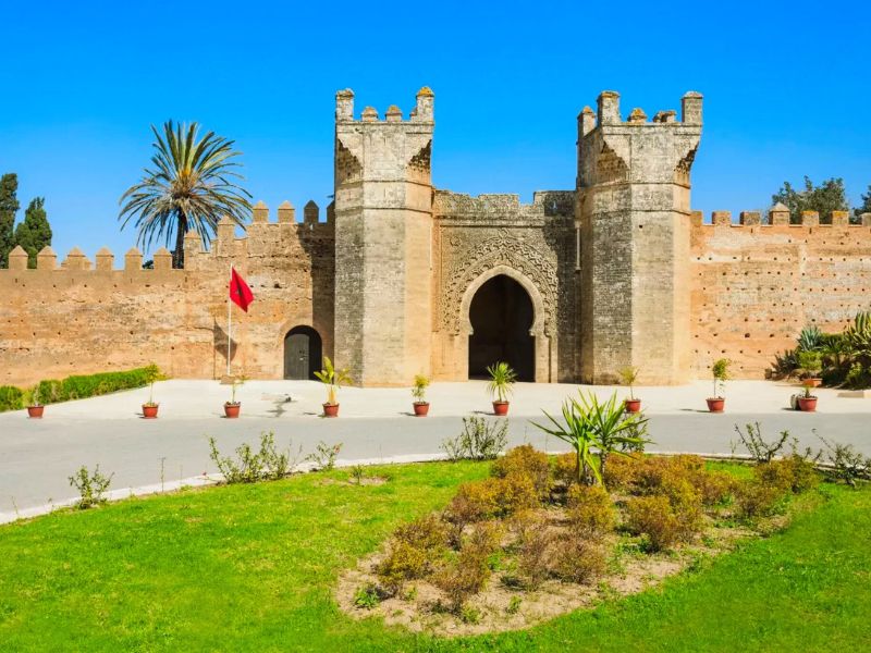 View of the entrance gate to the ancient Chellah site in Rabat, Morocco, featuring a tall stone structure with two towers, decorative archways, and palm trees in the background.