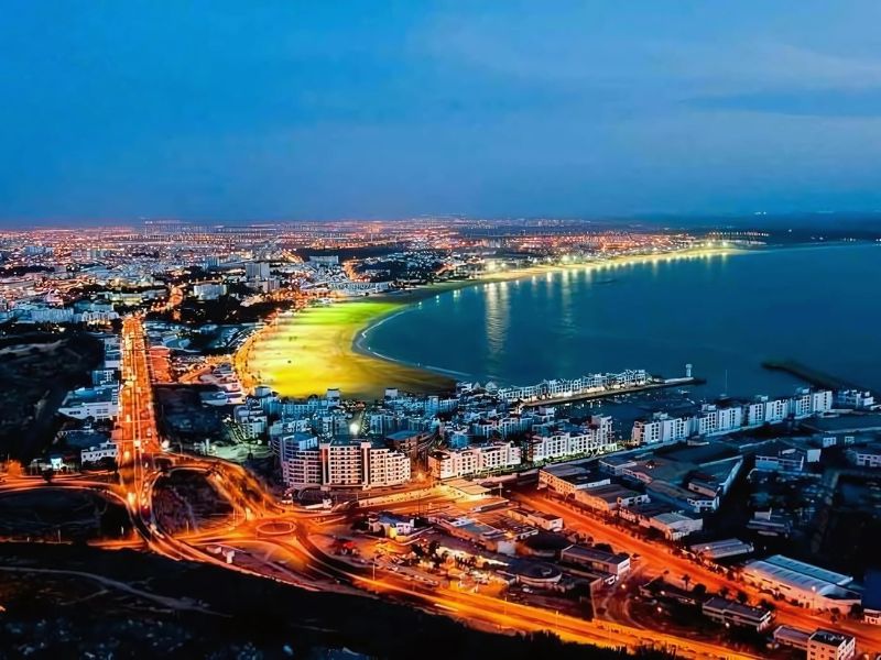 Aerial view of Agadir at night, showcasing the city's coastline, beach, and illuminated buildings along the shore.