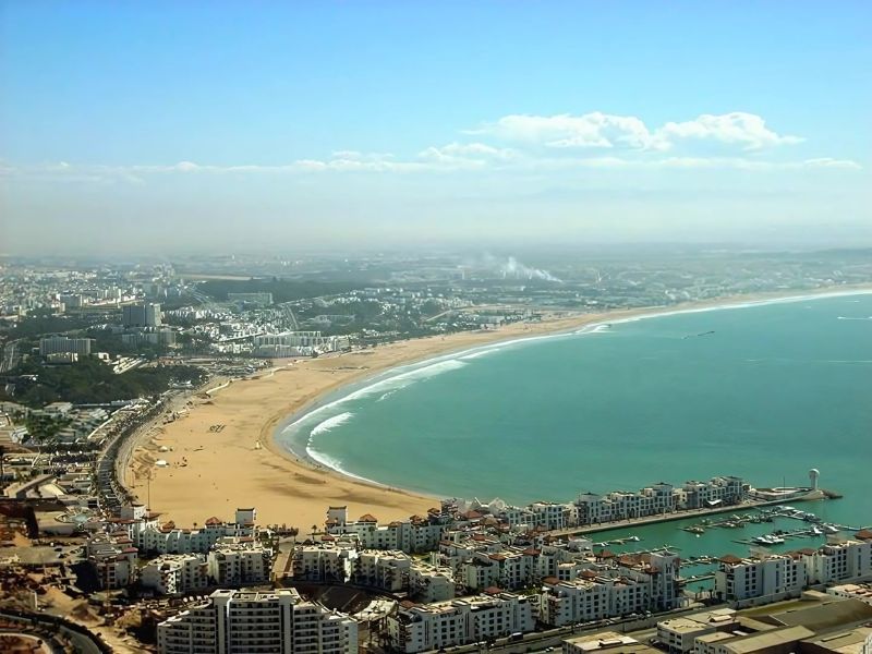 A panoramic view of Agadir, Morocco, showcasing the city along the Atlantic coast, with sandy beaches and modern buildings visible under a clear blue sky.