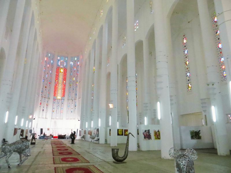 Interior view of the Casablanca Cathedral, showcasing its high vaulted ceiling, ornate stained-glass windows, and white pillars. The space includes decorative artworks and sculptures.