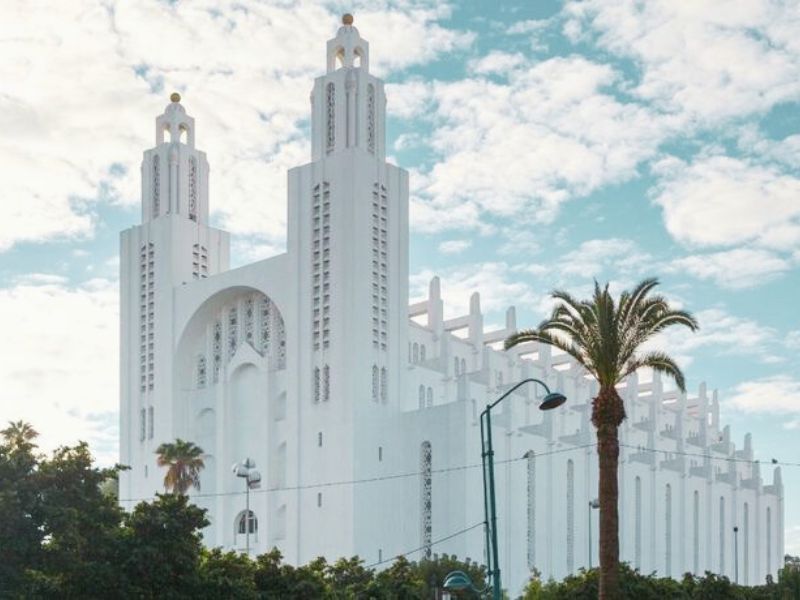 Exterior view of the Casablanca Cathedral, showcasing its white Art Deco and Neo-Gothic architecture, twin towers, and surrounding palm trees against a blue sky.
