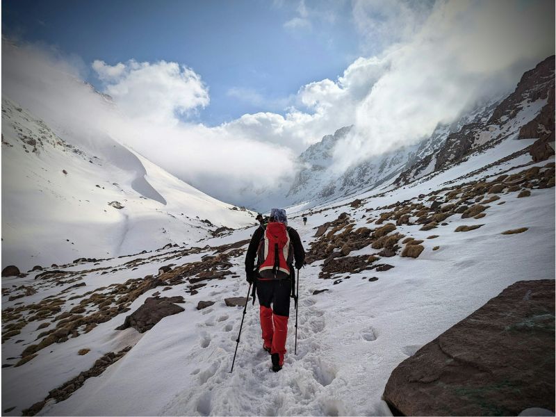 A hiker trekking through snow covered terrain towards Mount Toubkal, surrounded by dramatic mountain scenery and cloudy skies.
