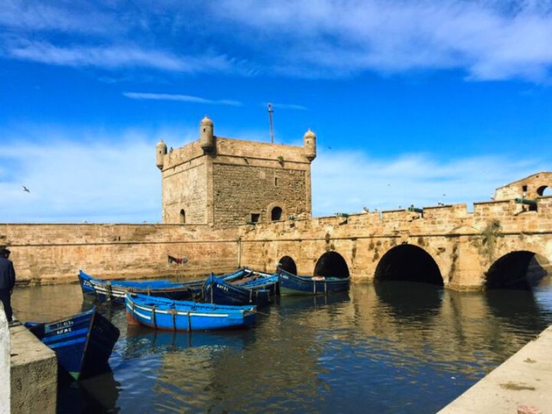 The Skalla de la Ville, an 18th-century fortification in Essaouira, Morocco, with stone walls and towers, surrounded by blue fishing boats on the water under a clear sky.