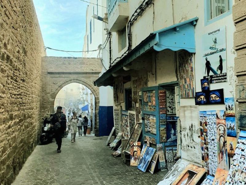 Narrow street in Essaouira's Medina, lined with whitewashed buildings and blue accents, showcasing local artwork and a bustling atmosphere.