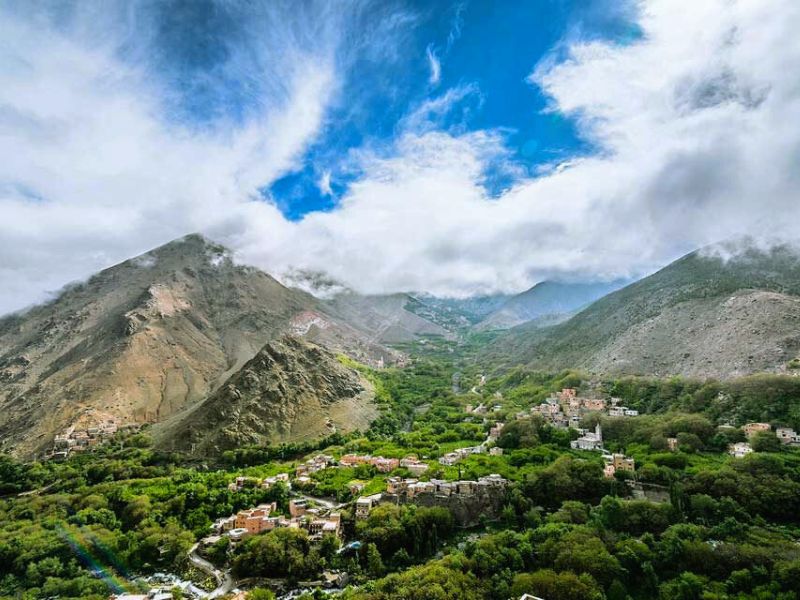 A panoramic view of Imlil village nestled in the Atlas Mountains, featuring lush greenery, surrounding peaks, and dramatic clouds in the sky.
