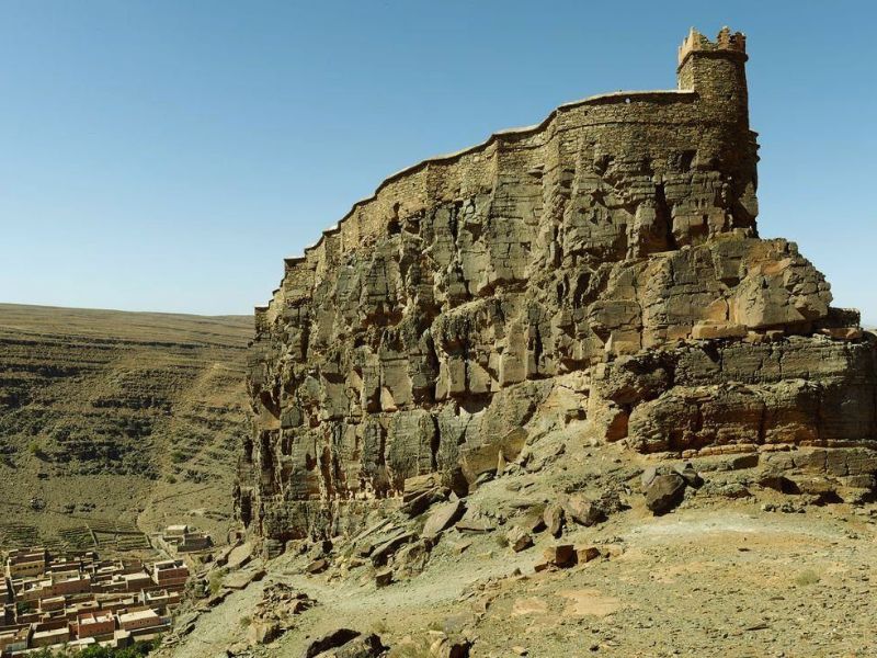 A historic granary known as Igoudar, located on a rocky cliff with a strong structure, overlooking a nearby village in a mountainous landscape.