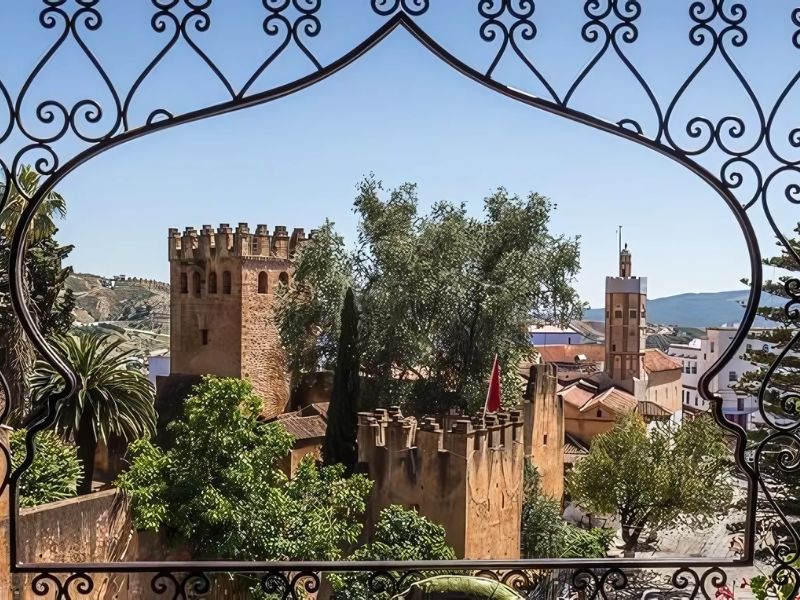 View of the Kasbah of Chefchaouen framed by intricate ironwork, showcasing the fortress walls, lush greenery, and buildings of the blue city under a clear sky.
