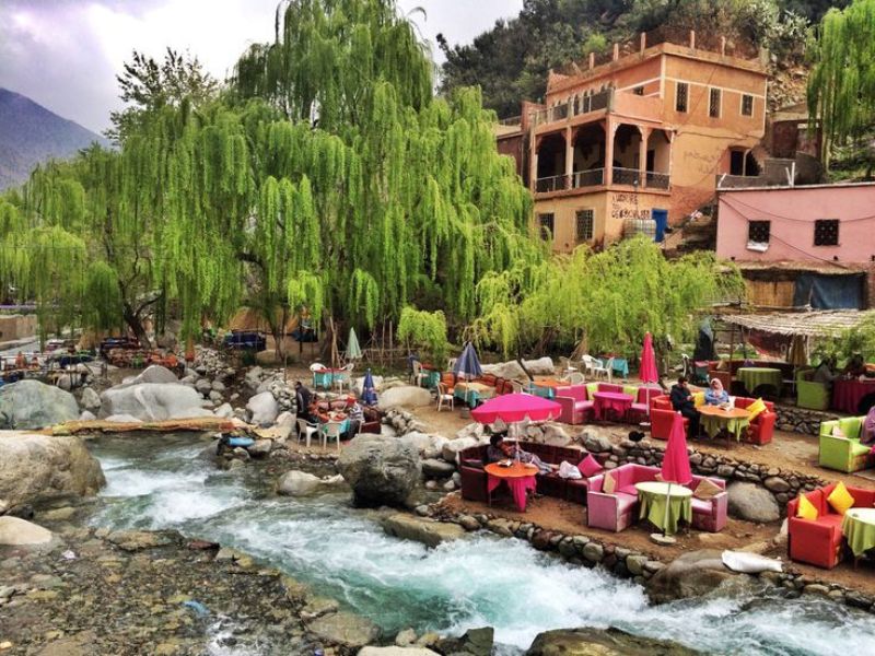 Scenic view of a riverside café in Ourika Valley, featuring colorful umbrellas and seating areas surrounded by lush greenery and flowing water.