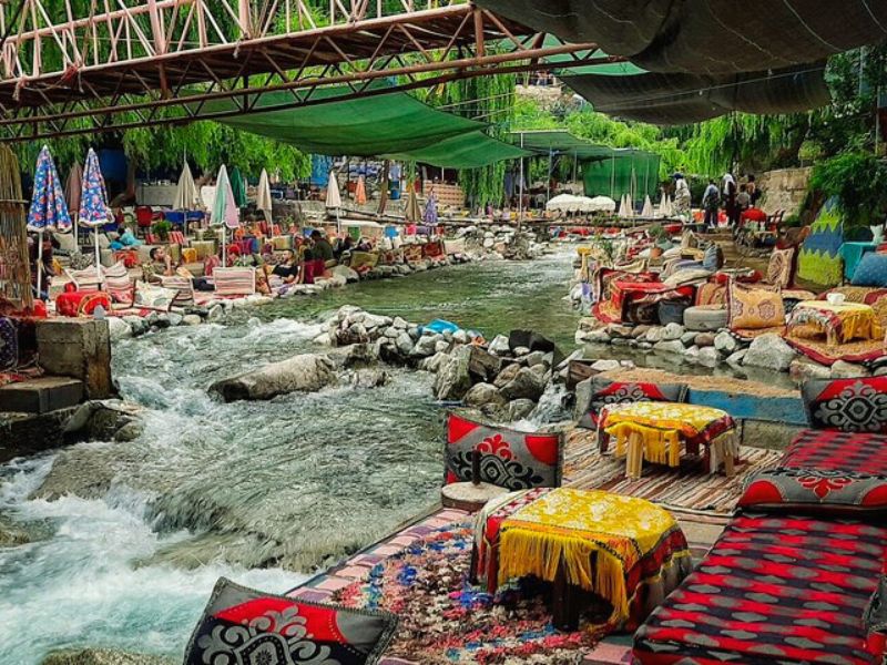 A vibrant seating area along the banks of a river in the Ourika Valley, featuring colorful cushions and traditional Moroccan decor, surrounded by greenery and glimpses of visitors enjoying the scenery.