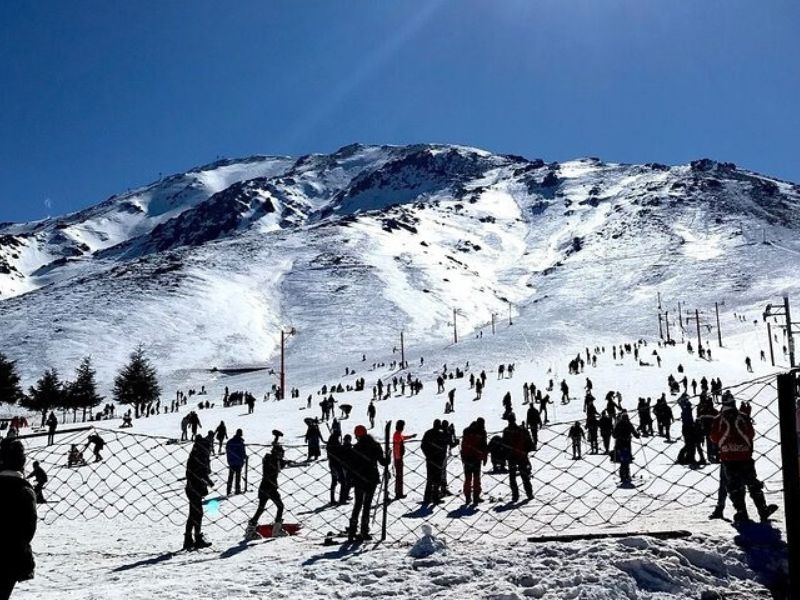 A snowy ski resort in Oukaimden, featuring numerous people skiing and snowboarding on the slopes, surrounded by towering mountains under a clear blue sky.