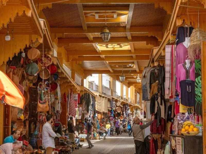 A vibrant market scene in the Medina of Rabat, showcasing colorful textiles, handcrafted goods, and traditional Moroccan wares, with people strolling and shopping in a bustling alley.