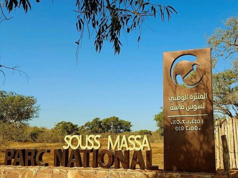 Sign marking the entrance to Souss Massa National Park, featuring the park's name in Arabic French and Amazigh against a natural background.