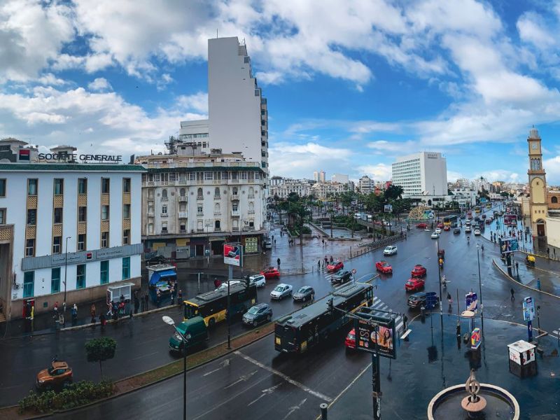 A bustling street view of Casablanca, Morocco, showcasing a mix of modern and colonial architecture, with various vehicles on the wet road and a blue sky with scattered clouds.