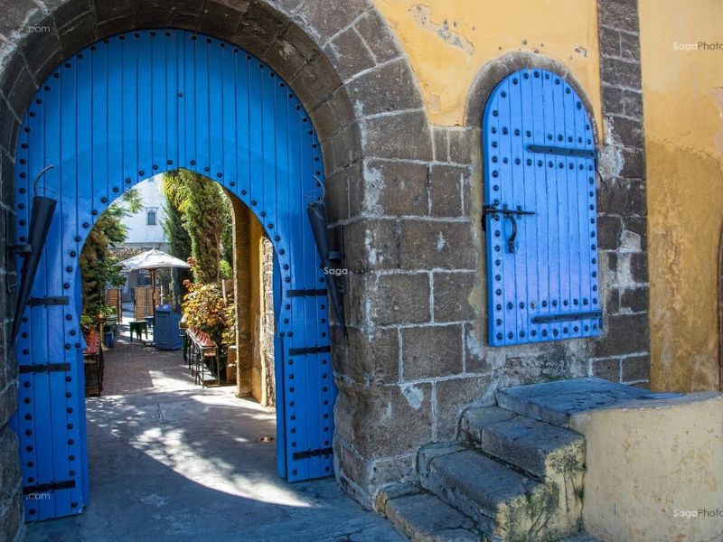 A vibrant blue wooden door with intricate detailing, set in a stone archway, leading to a courtyard adorned with greenery and outdoor seating, highlighting traditional Moroccan architecture.