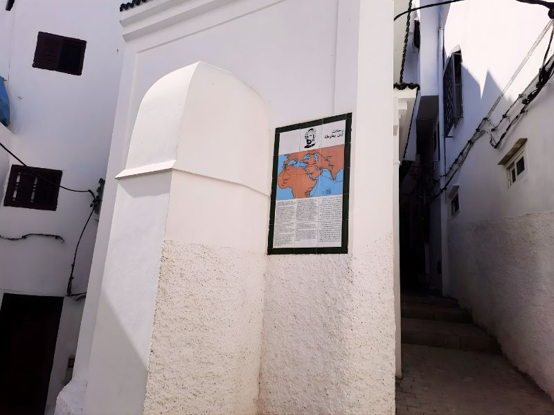 An image of a narrow street in Tangier featuring a white wall with a rounded top and a display board that includes a map of the world and descriptive text about Ibn Battuta.
