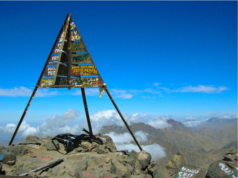 Summit marker at Mount Toubkal, the highest peak in North Africa, with a clear blue sky and mountains in the background.