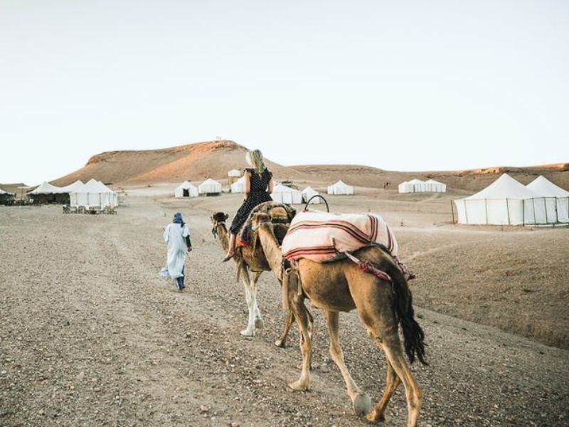 A woman riding a camel in the Agafay Desert, accompanied by a guide walking alongside, with desert tents in the background.