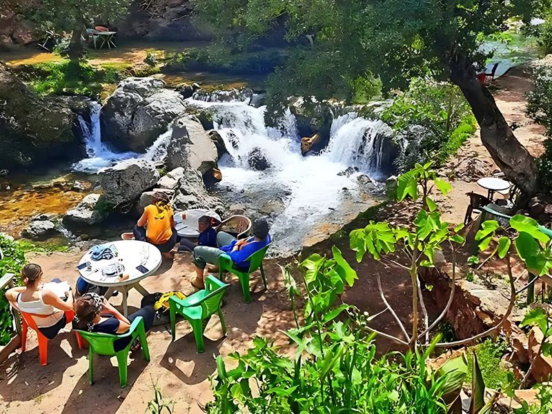 A group of people relaxing around a table near picturesque waterfalls in Akchour, Morocco, surrounded by lush greenery.