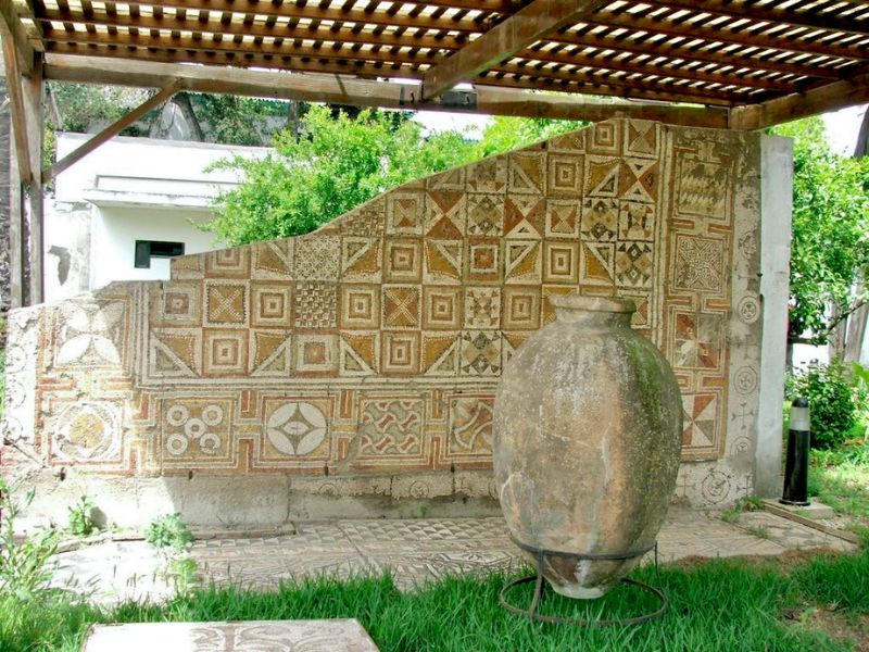 An outdoor display at the Archaeological Museum of Tetouan featuring a patterned wall and an ancient pottery vessel, surrounded by greenery.