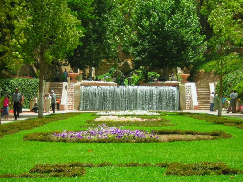 A lush green park in Beni Mellal, Morocco, featuring colorful flower beds and a cascading waterfall, with people walking along the pathways.