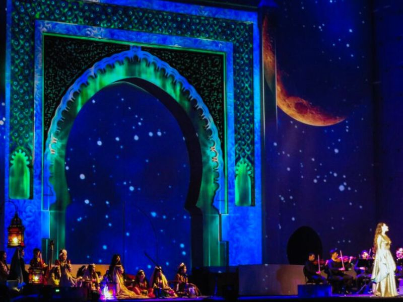 A performance scene from the Fes World Sacred Music Festival featuring a singer in a flowing dress on stage, with a backdrop of colorful lights and stars, highlighting the festival atmosphere.