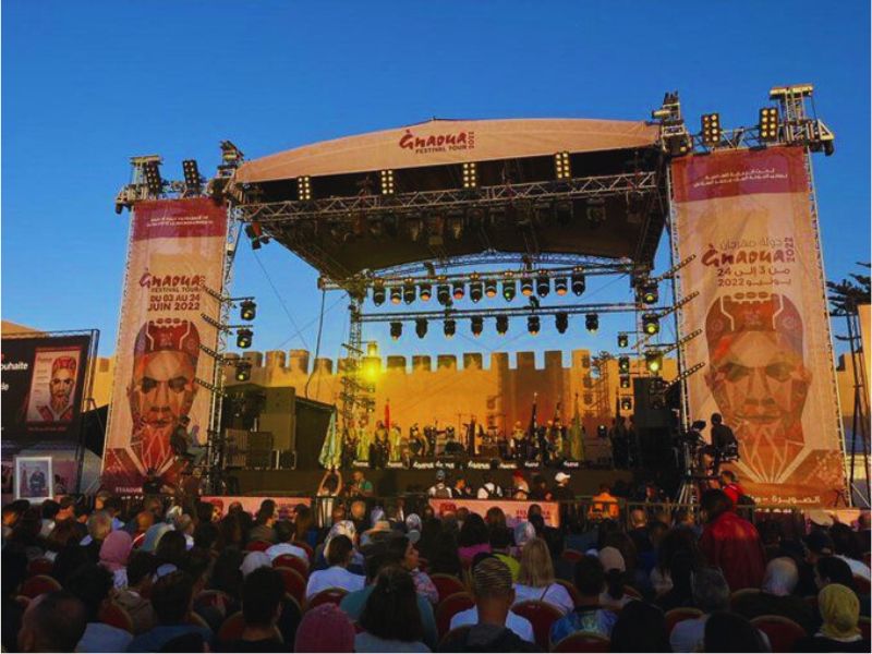 A stage set for the Gnaoua and World Music Festival in Essaouira, featuring performers and a diverse audience gathered under a clear blue sky.