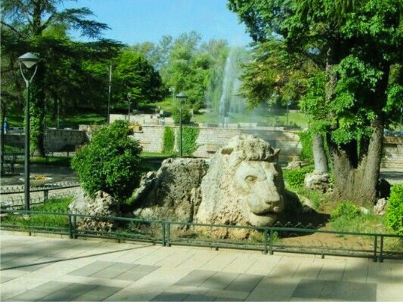 A large stone lion sculpture in a park setting in Ifrane, Morocco, surrounded by greenery and trees.