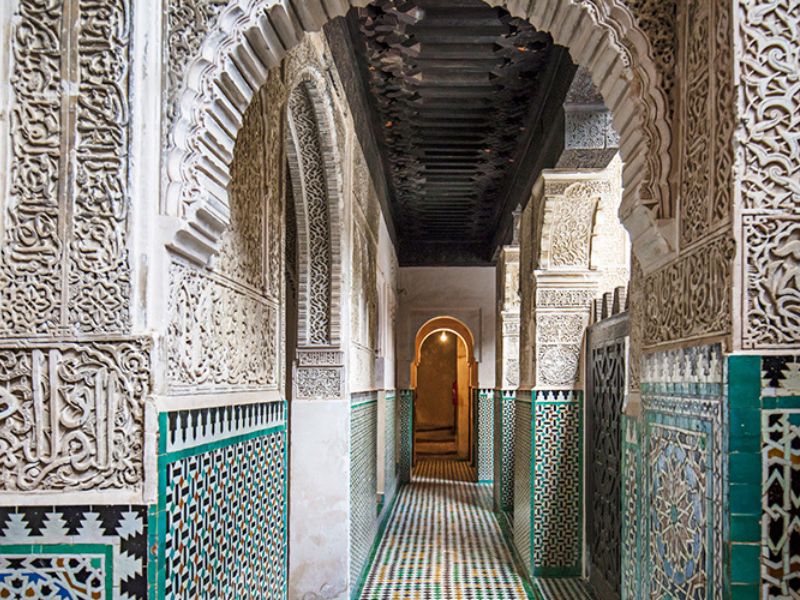 Interior corridor of the Medersa Bou Inania in Fez, showcasing intricate carved plasterwork, colorful Zellij tile patterns, and a wooden ceiling.