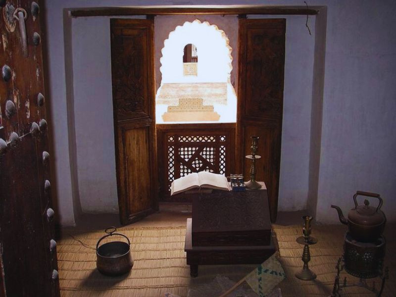 Interior view of a traditional room in the Medersa Ben Youssef, featuring wooden doors, a decorative window, an open book on a wooden stand, and traditional Moroccan artifacts.