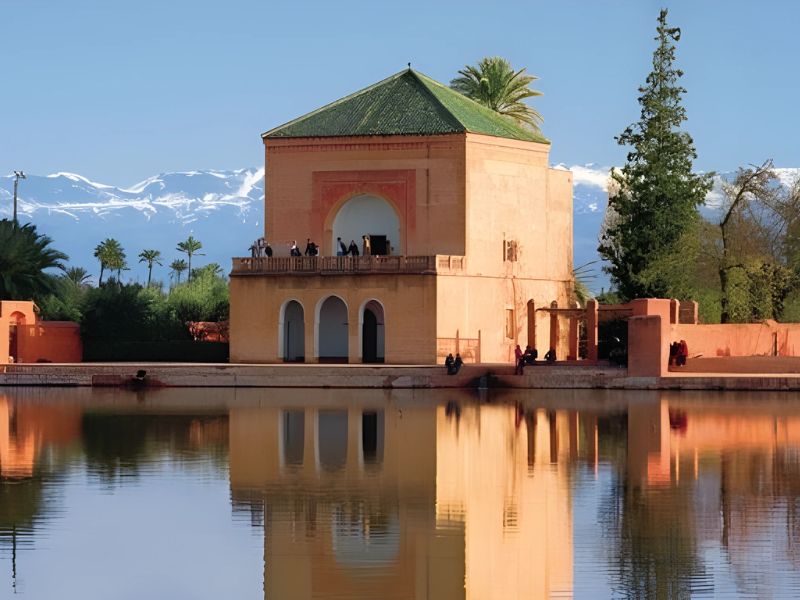 Central pavilion of the Menara Gardens in Marrakesh, surrounded by a tranquil pond, with lush greenery and the Atlas Mountains in the background.
