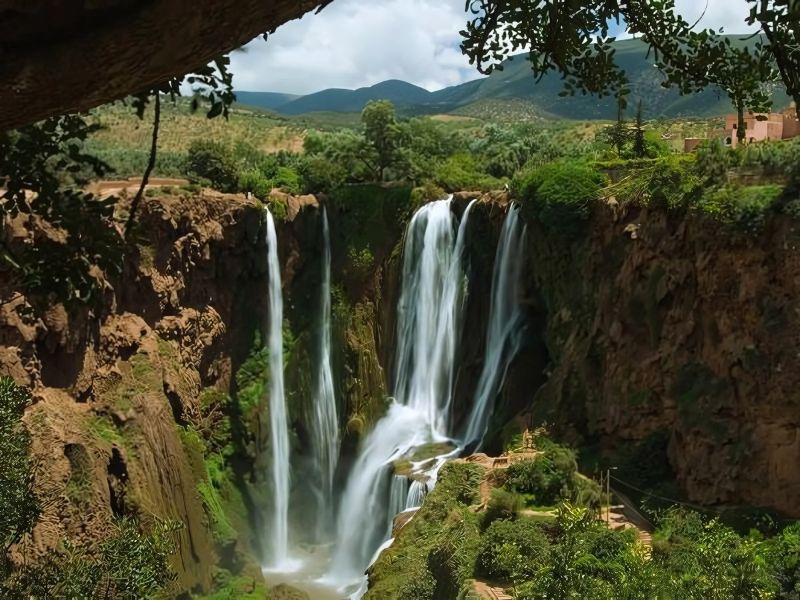 A stunning view of the Ouzoud Waterfalls in Morocco, featuring three cascading falls that plunge into a lush green valley surrounded by trees and mountains.