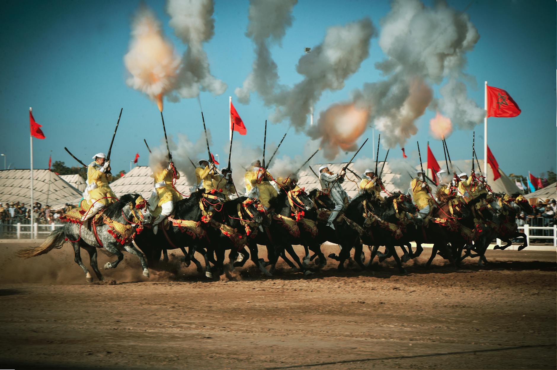 A thrilling Tbourida performance featuring a synchronized charge of riders on horseback, firing antique rifles amidst clouds of smoke and vibrant flags against a clear blue sky.