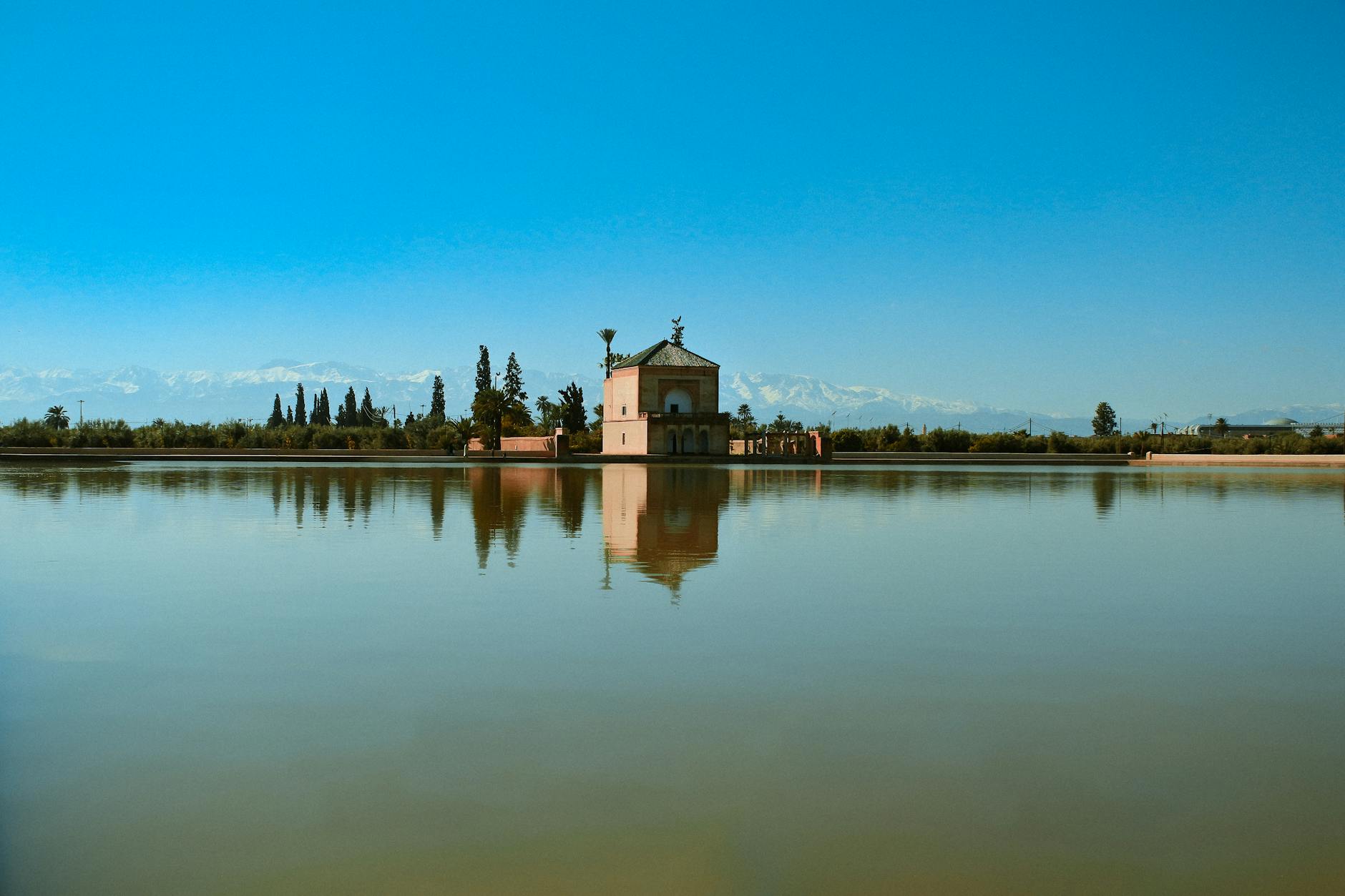 A tranquil view of the Menara Gardens in Marrakesh, showcasing the central pond and a historic pavilion, with the Atlas Mountains in the background under a clear blue sky.