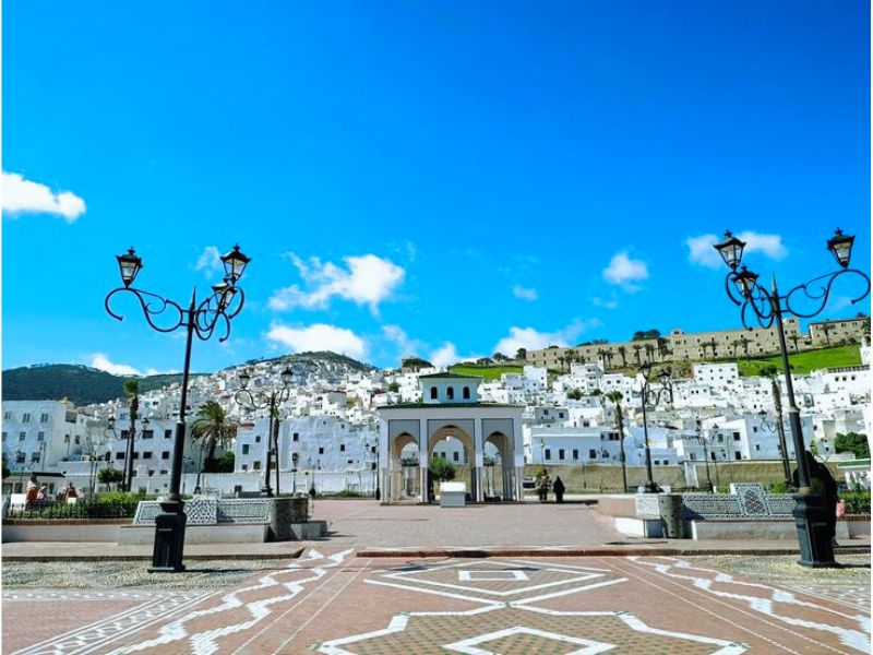 A view of the town of Tetouan, featuring whitewashed buildings at the foot of a mountain under a bright blue sky.