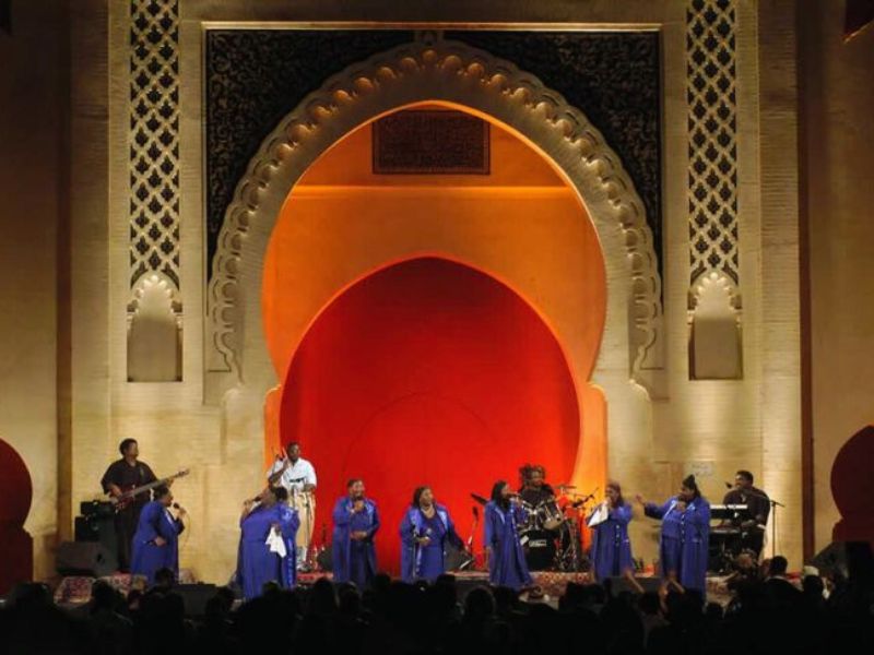 A live performance at the Fez Festival of World Sacred Music showcasing musicians in blue robes on stage, with an elaborate architectural backdrop.