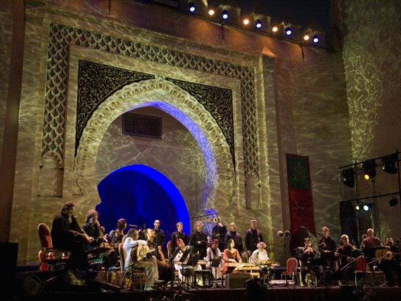 A performance at the Fes World Sacred Music Festival featuring a diverse group of musicians on stage in a historic venue, illuminated by colorful lights.