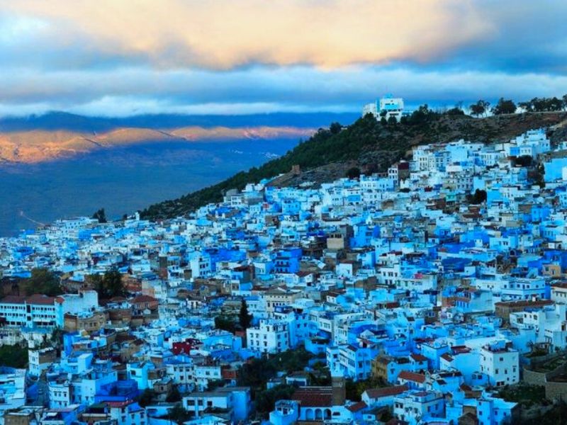 A panoramic view of the blue-washed city of Chefchaouen, set against a mountainous backdrop with clouds and light illuminating the scene.