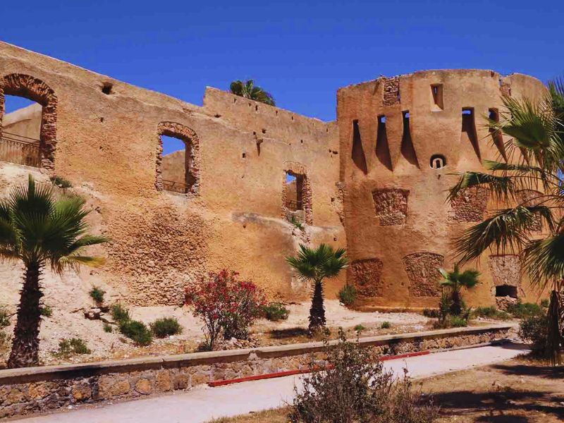 Ruins of a fortified structure in Azemmour, Morocco, featuring palm trees and vibrant vegetation in the foreground against a clear blue sky.