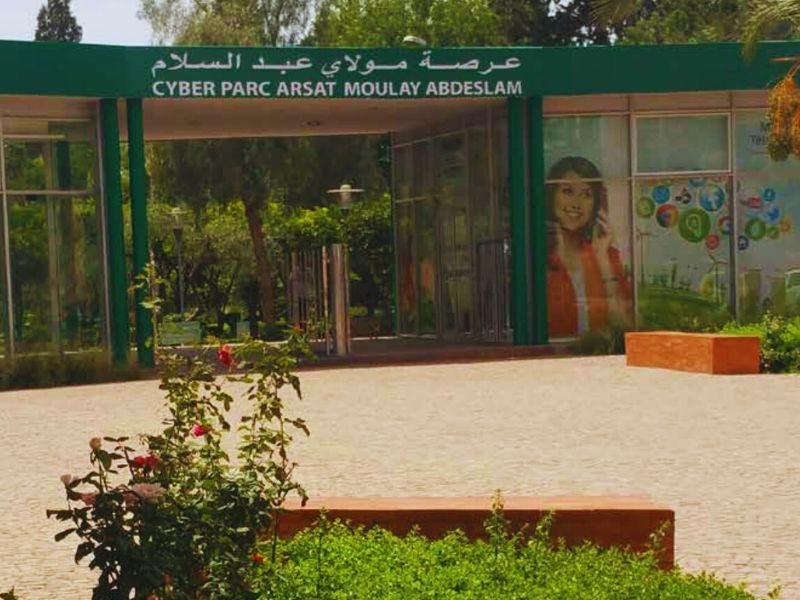 Entrance to Arsat Moulay Abdessalam, also known as Cyber Parc, featuring a green sign with the park's name in Arabic and English, surrounded by lush greenery and colorful flowerbeds.